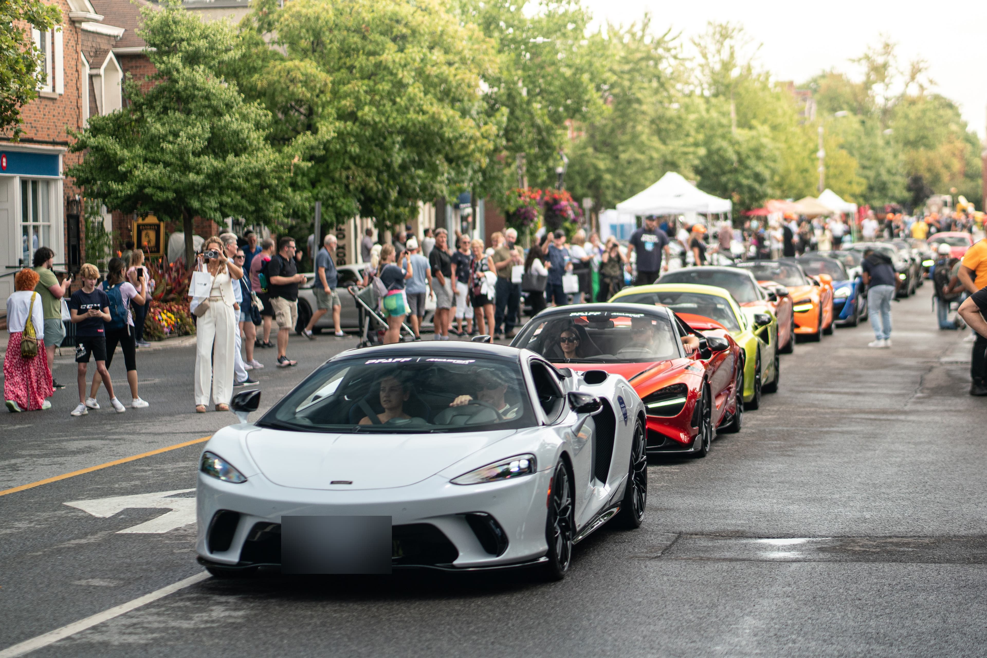 White McLaren GT leads a supercar parade past crowds on a village street