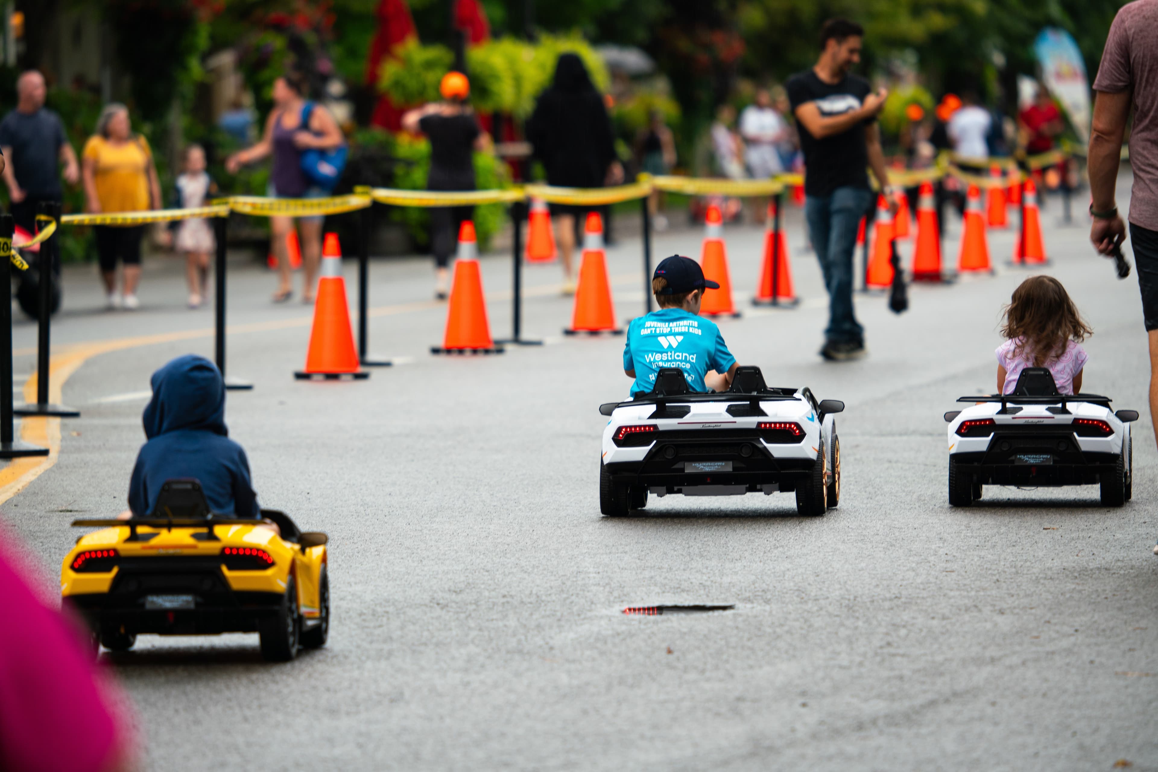 Children drive toy Lamborghini ride-on cars along a coned-off street