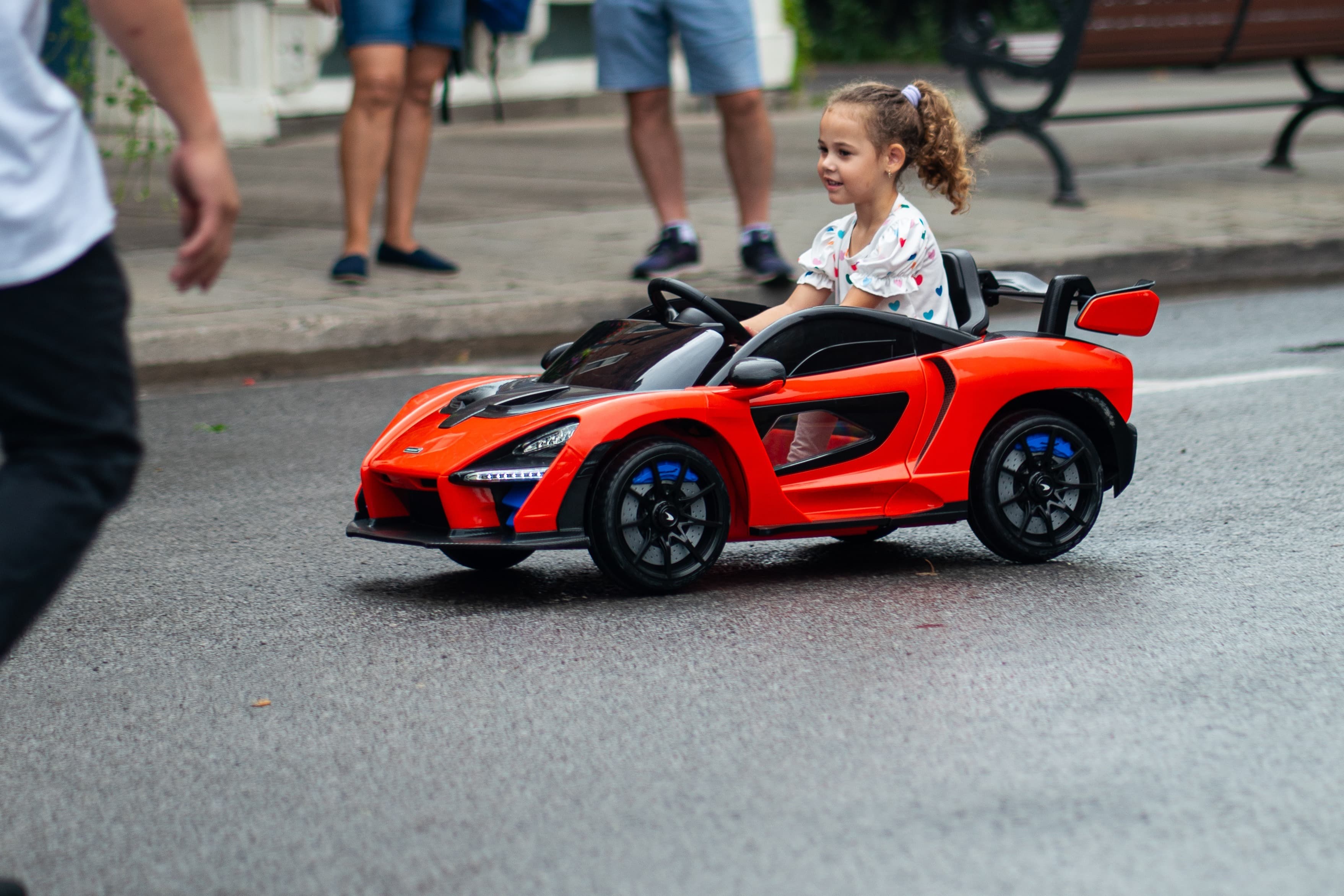 Young girl drives a red McLaren ride-on toy car along a street
